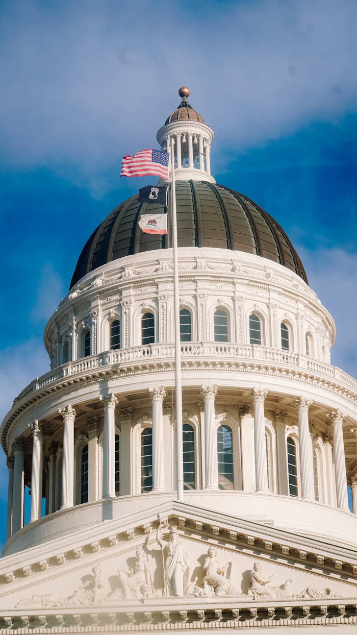 Close-up of the California State Capitol Building dome with flags, clear blue sky.